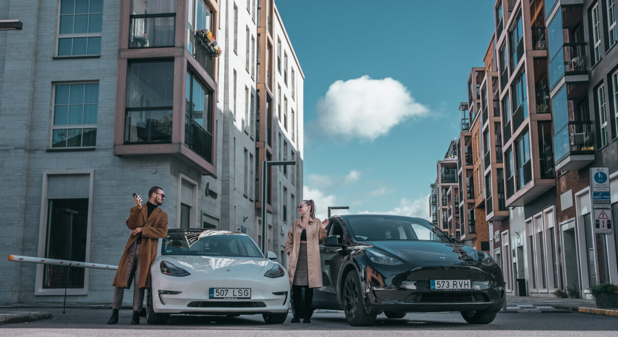 Man and a woman standing next to two Teslas. One white Model 3 Tesla and one Model Y Tesla. In a city centre. Holding pones.