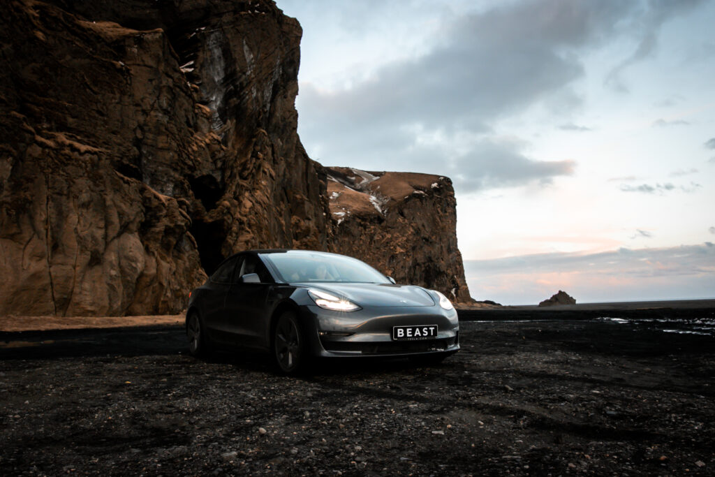 Black Model 3 Tesla in Iceland in the background there are mountains and it's on the black sand beach.